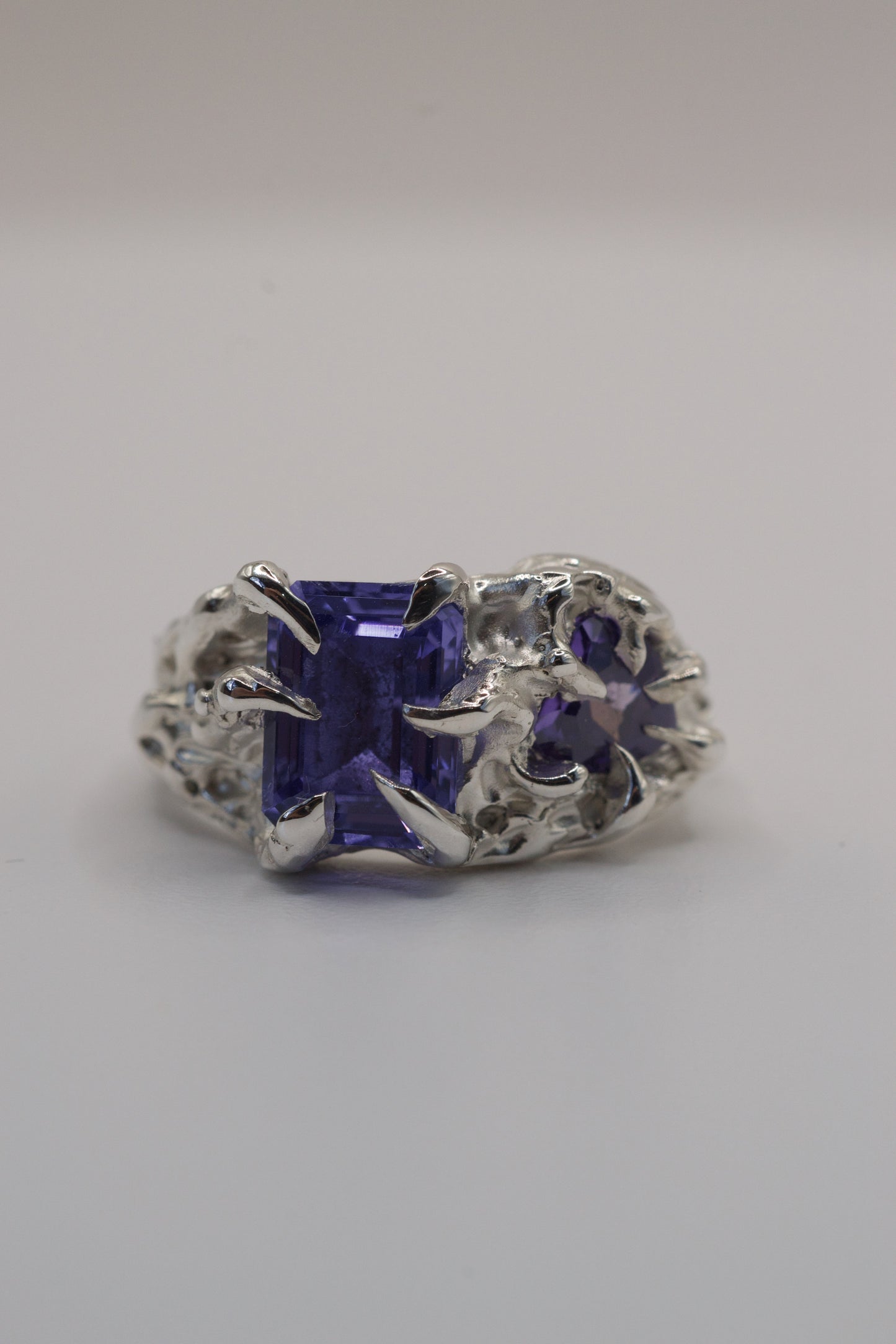 Front view of a handmade silver ring with a deep purple rectangular gemstone held in a gothic-style claw setting. The design is textured and organic, created using the lost wax casting method.