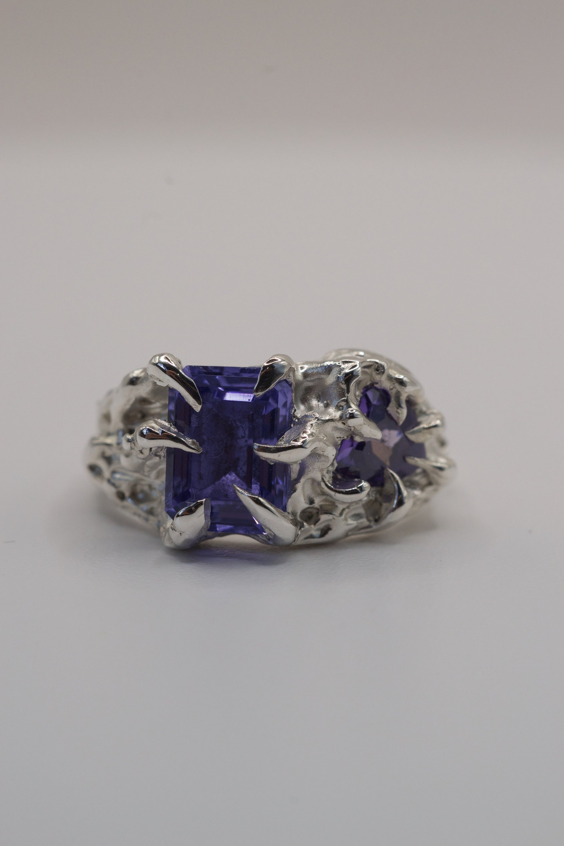 Front view of a handmade silver ring with a deep purple rectangular gemstone held in a gothic-style claw setting. The design is textured and organic, created using the lost wax casting method.