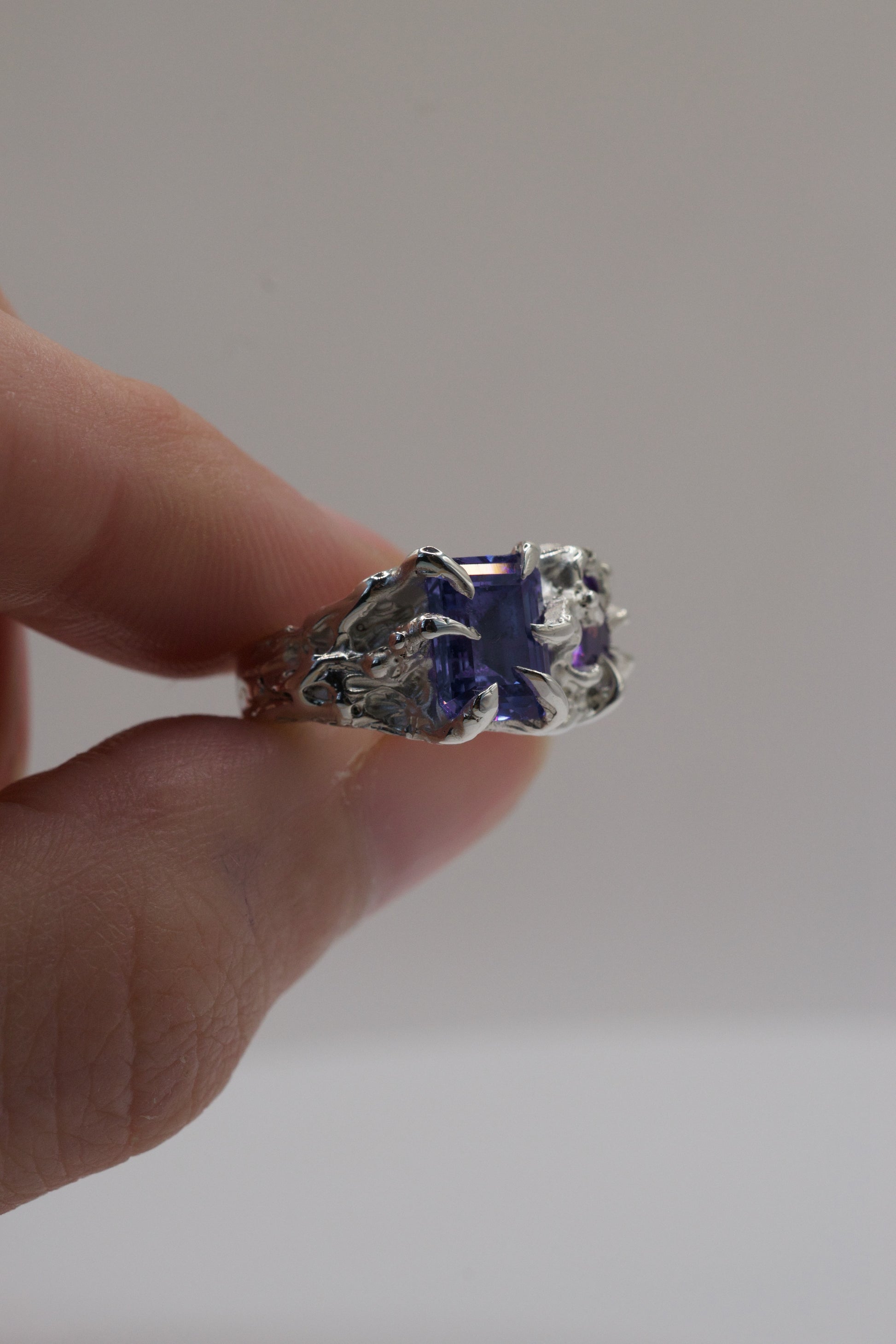 Silver ring with a purple gemstone held by a hand against a plain background